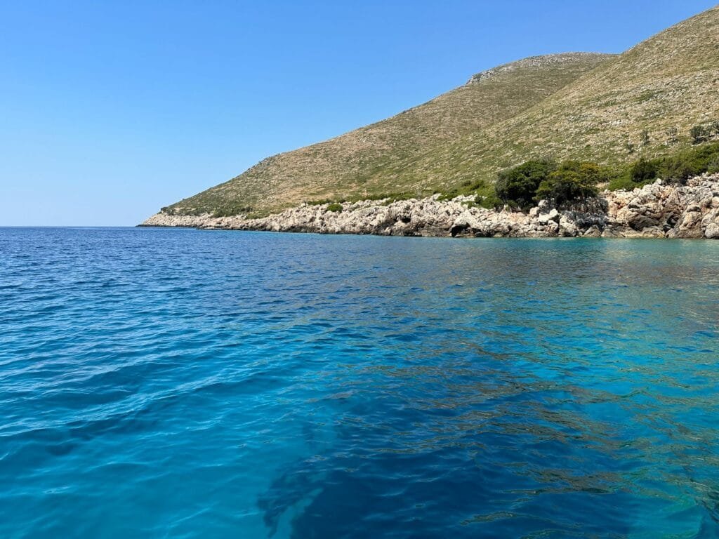 Stunning view of Grama Bay, Albania, showcasing crystal-clear turquoise waters meeting the rugged rocky coastline and green hills. A hidden paradise on the Ionian Sea, Grama Bay is a top destination for boat tours, snorkeling, and exploring Albania’s untouched nature.