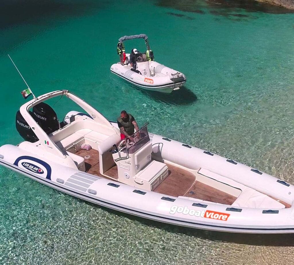 Two luxury inflatable boats anchored in the crystal-clear waters of Grama Bay, Albania. The "GoBoat Vlore" vessel sits near the shore, showcasing the pristine beach and turquoise sea, making it an ideal destination for boat tours and summer adventures along the Albanian Riviera.