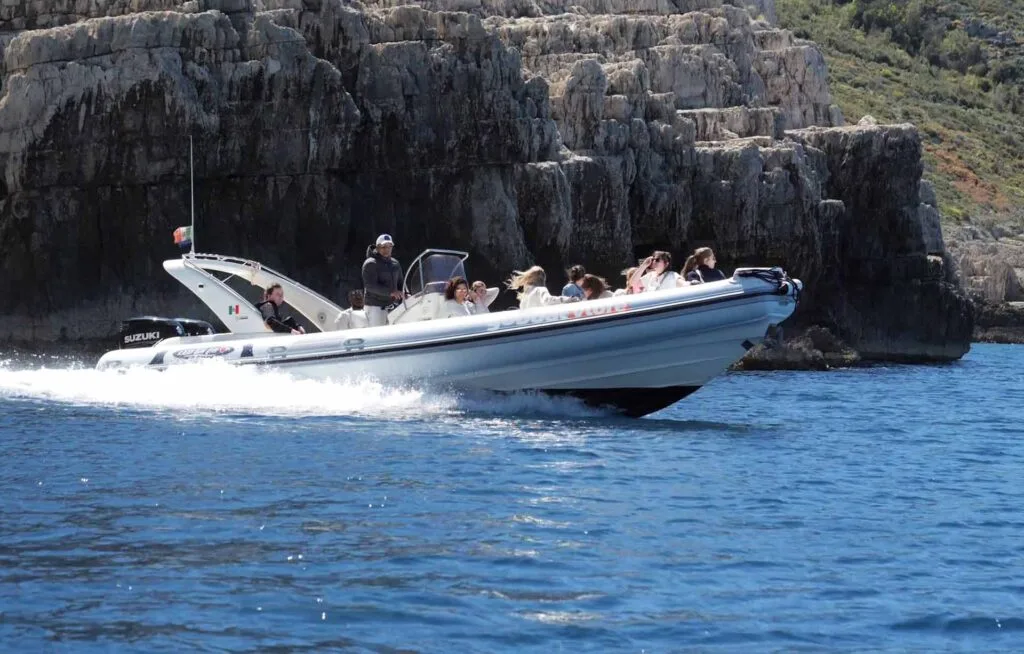A speedboat full of excited travelers cruising away from Haxhi Ali Cave, Albania. The boat glides over the crystal-clear waters of the Adriatic Sea, passing by dramatic limestone cliffs.
