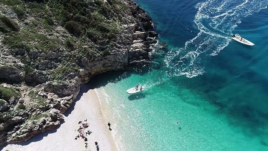 A stunning aerial view of a hidden turquoise beach in Albania, where a speedboat carves a circular wake near the shore. Tourists unwind on the soft white sand, while another boat floats in the shallow, crystal-clear waters. Beneath the surface, mesmerizing underwater rock formations highlight the unspoiled beauty of the Karaburun Peninsula